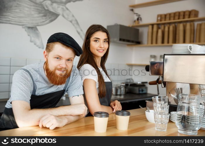 Coffee Business Concept - Positive young bearded man and beautiful attractive lady barista couple in apron looking at camera while standing at bar Couter ready to give Coffee Service at the modern coffee shop