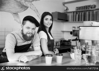 Coffee Business Concept - Positive young bearded man and beautiful attractive lady barista couple in apron looking at camera while standing at bar Counter.. Coffee Business Concept - Positive young bearded man and beautiful attractive lady barista couple in apron looking at camera while standing at bar Counter