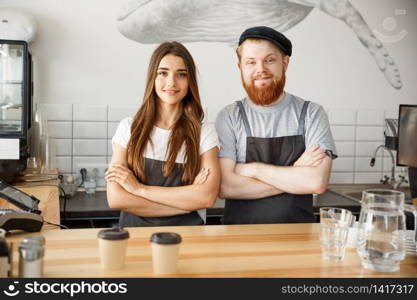 Coffee Business Concept - Positive young bearded man and beautiful attractive lady barista couple in apron looking at camera while standing at bar Couter ready to give Coffee Service at the modern coffee shop