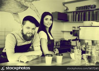 Coffee Business Concept - Positive young bearded man and beautiful attractive lady barista couple in apron.. Coffee Business Concept - Positive young bearded man and beautiful attractive lady barista couple in apron looking at camera while standing at bar Counter