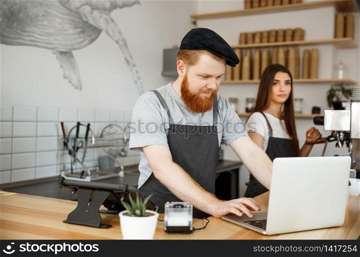 Coffee Business Concept - Young handsome bearded bartende, barista or manager working and planing in laptop at modern coffee shop.