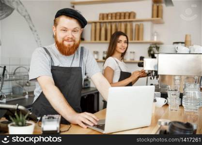 Coffee Business Concept - Young handsome bearded bartende, barista or manager working and planing in laptop at modern coffee shop.