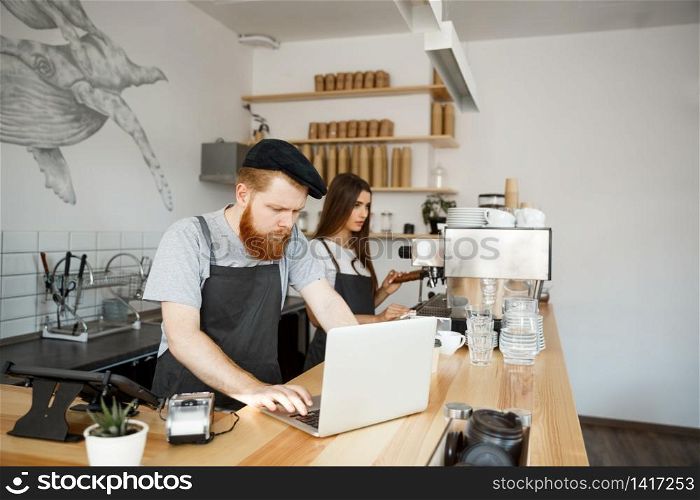 Coffee Business Concept - Young handsome bearded bartender, barista or manager working and planing in laptop at modern coffee shop. Coffee Business Concept - Young handsome bearded bartender, barista or manager working and planing in laptop at modern coffee shop.