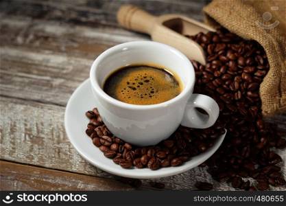 Coffee cup and roasted coffee beans in a burlap sack on a rustic wooden table on rusty vintage background.