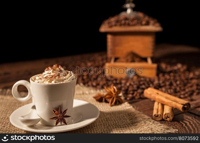 Coffee cup with whipped cream, cocoa powder and star anise with coffee grinder on background