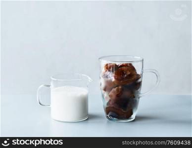 Coffee ice cubes in glass mug and little jug with cream or vegan milk on grey table at wall background. Refreshing summer drinks making ingredients