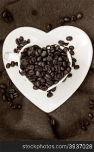 Coffee klatsch java concept. Heart shaped white cup filled with roasted coffee beans on brown cloth background