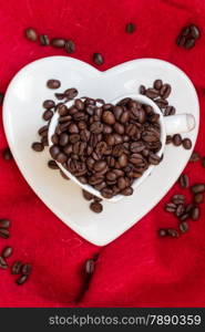 Coffee klatsch java concept. Heart shaped white cup filled with roasted coffee beans on red cloth background