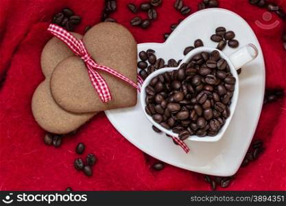 Coffee love concept. Coffee beans in heart shaped white cup and sweet cookie gingerbread with ribbon on red cloth background. Top view