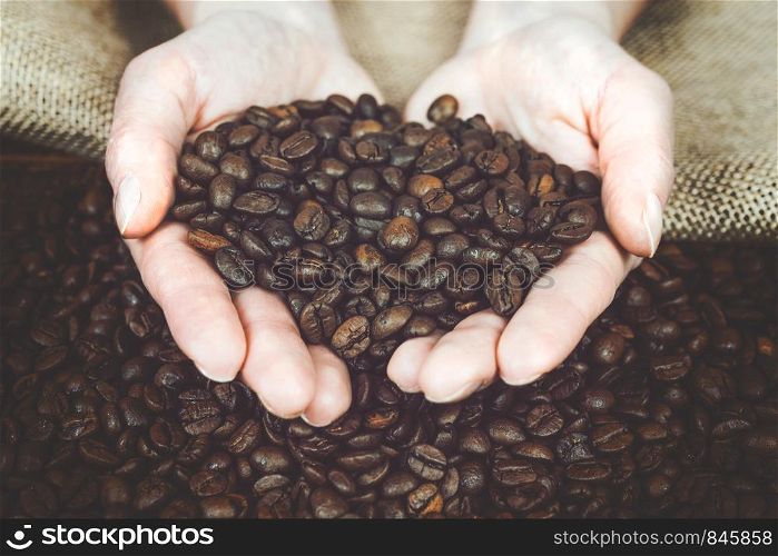 Coffee production concept - woman is holding fresh roasted beans in her hands over a wooden desk and a burlap sack (vintage effect).