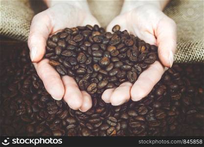 Coffee production concept - woman is holding fresh roasted beans in her hands over a wooden desk and a burlap sack (vintage effect).