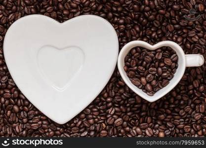 Coffee time. White cup and saucer in heart shape on roasted coffee beans background. Top view