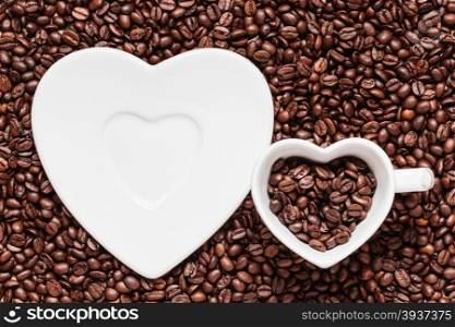 Coffee time. White cup and saucer in shape of heart on roasted coffee beans background. Top view