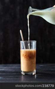 Coffee with milk. On a black wooden table, milk is poured into a glass of coffee.. In freshly prepared coffee in a transparent glass pour milk on a black wooden table. Delicious morning drink.