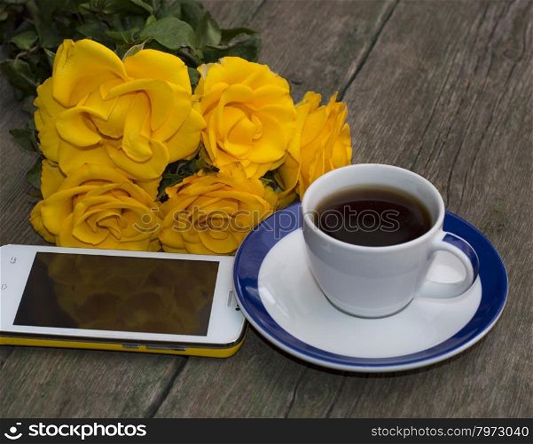 coffee, yellow roses and the mobile phone on a wooden table