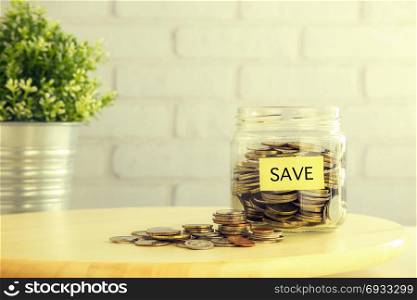 Coins in saving glass jar and on wooden table with yellow SAVE tag, bricks and plant pot on background vintage retro style. Save money for future financial planning, investment, education or retirement funds.