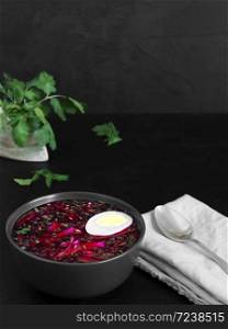 Cold beetroot soup in a black ceramic plate with half a boiled egg. Summer cold soup. A bowl on a gray napkin, next to a spoon and sprigs of cilantro. Copy space. Black background. Healthy, natural food. Close-up.