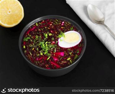 Cold beetroot soup in a black ceramic plate with half a boiled egg. Summer cold soup. Next to a gray napkin is a spoon and half a lemon. Black background. Healthy, natural food. Flat lay.. Cold beetroot soup in a black ceramic plate with half a boiled egg. Next to a gray napkin is a spoon and half a lemon. Black background. Healthy, natural food. Top view.