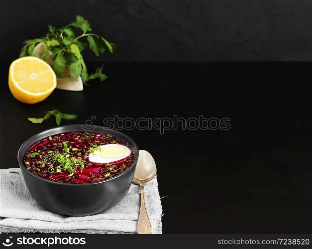 Cold beetroot soup in a black ceramic plate with half a boiled egg. Summer cold soup. A bowl on a gray napkin, next to a spoon, half a lemon and cilantro. Copy space. Black background. Healthy, natural food. Close-up.