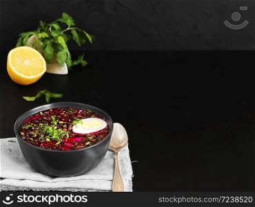 Cold beetroot soup in a black ceramic plate with half a boiled egg. Summer cold soup. A bowl on a gray napkin, next to a spoon, half a lemon and cilantro. Copy space. Black background. Healthy, natural food. Close-up.