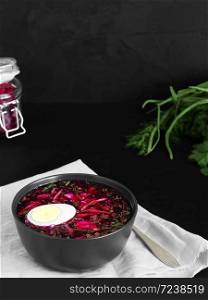 Cold borsch in a black ceramic plate with half a boiled egg. Summer cold soup. Bowl on a gray napkin, next to a spoon. In the background is a jar with slices of beets and fresh herbs. Black background. Close-up.
