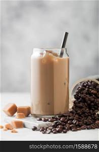 Cold iced coffee with milk in glass with jar of fresh coffee beans and salted caramel on light table background.