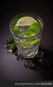 Cold refreshing summer lemonade mojito in a glass on a black concrete or stone background.. Refreshing cold Caipirinha with fresh lime slices and ice on a dark slate