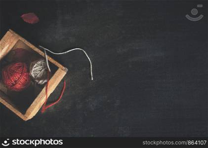 Cold season activities concept with thread balls in a wooden crate and red fallen leaves on a rustic black background. Leisure context. Knitting craft