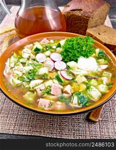 Cold soup okroshka from sausage, potato, egg, radish, cucumber, greens and kvass in a bowl, spoon, bread and jug with drink on wicker napkin on wooden board background
