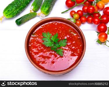 Cold tomato soup gazpacho from fresh tomato and vegetables in a brown round plate on a white wooden table, top view
