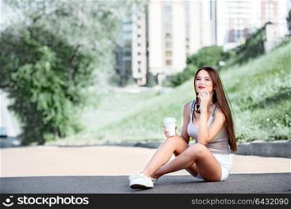 Collage of a beautiful young woman with music headphones around her neck, drinking coffee from a takeaway coffee cup and sitting on the road near a separating strip.