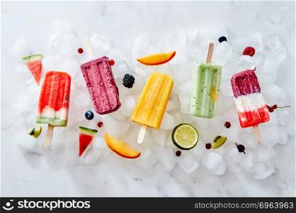 Collection of colorful tasty homemade ice cream lolly on a stick with juicy berries and pieces of various fruits on ice cubes. Flat lay. Different ice cream on a stick and different pieces of fruit and berries on ice cubes. Flat lay