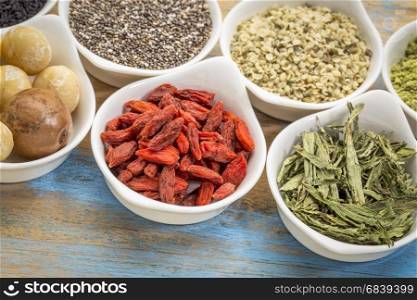 collection of superfoods in small ceramic bowls against rustic wood: macadamia nuts, hemp seed hearts, goji berry, stevia herb and chia seeds