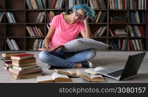 College female student sitting on the floor cross legged with a book, tired and exhausted preparing for exam. Hipster girl in trendy eyeglasses surronded by books studying hard in university library, reading a book and making notes on laptop.