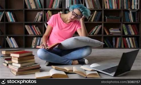 College female student sitting on the floor cross legged with a book, tired and exhausted preparing for exam. Hipster girl in trendy eyeglasses surronded by books studying hard in university library, reading a book and making notes on laptop.
