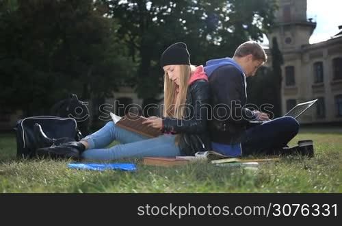 College students studying together near campus. Couple of students sitting back to back on the grass and studying. Woman making notes in textbook with pen. Man typing on laptop.