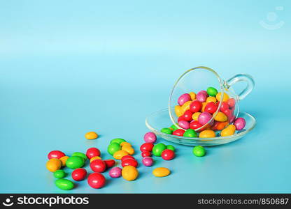 Colored glazed candy beans. Glass mug on saucer and scattered colorful button-shaped chocolates on blue background with copy space. Concept sweet tea party. Template for design, text.. Colored glazed candy beans. Glass mug on saucer and scattered colorful button-shaped chocolates on blue background with copy space. Concept sweet tea party. Template for design, text