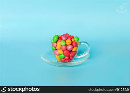 Colored glazed candy beans. Glass mug on saucer filled with colorful button-shaped chocolates on blue background with copy space. Concept sweet tea party. Template for design, text.. Colored glazed candy beans. Glass mug on saucer filled with colorful button-shaped chocolates on blue background with copy space. Concept sweet tea party. Template for design, text