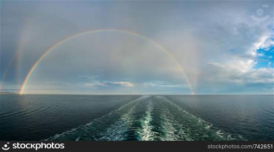 Colorful and dramatic double rainbow forms a full arch over the wake of a departing cruise ship at sea. Dramatic double rainbow forms over the wake of a cruise ship at sea
