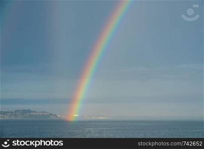 Colorful and dramatic double rainbow forms by the coastline and seen from a ship in the ocean. Dramatic rainbow forms over the wake of a cruise ship at sea
