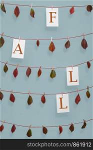 Colorful autumn frame with the word fall written on paper sheets and red dried leaves hanging on strings with wooden clips, on a blue wall.