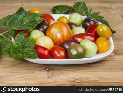 Colorful cherry tomatoes on white plate with green plant stems and leaves on bamboo wood
