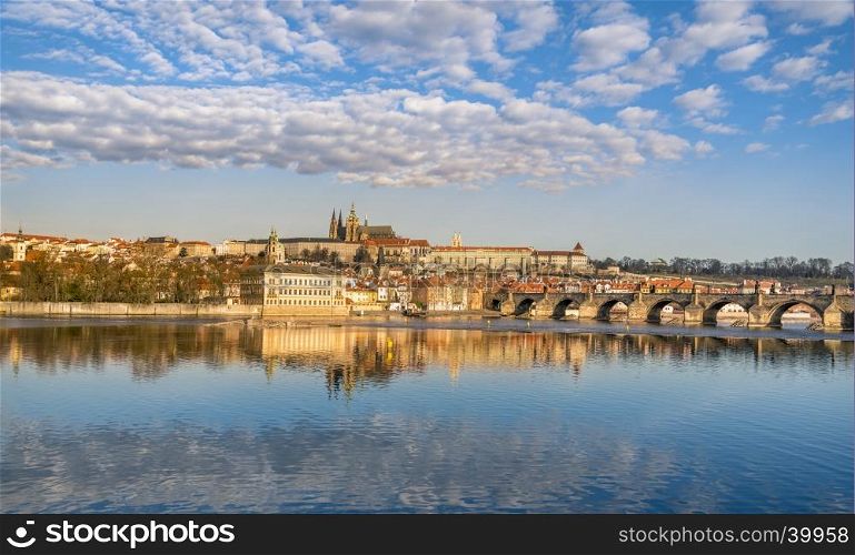 Colorful cityscape with the Charles Bridge, the Vltava river and Prague's buildings, under a blue sky, dotted with clouds and reflected in the water.