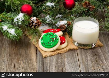 Colorful cookies and milk with snow covered evergreen branches, ornaments on rustic wood.