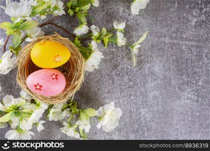 Colorful easter eggs in straw nest and blossoming branch with white flowers on gray concrete background. Top view, flat lay with copy space for text. Colorful easter eggs in nest and blossoming branch on grey concrete background
