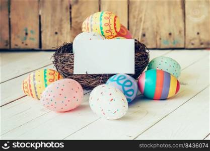 Colorful easter eggs in the nest and paper card on wood table background.