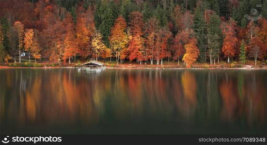 Colorful fall landscape with autumn forest and a wooden cottage reflected on the water of the Alpsee lake, near Fussen town, south Germany.