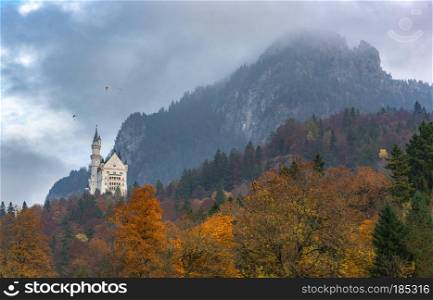 Colorful fall landscape with autumn forest, mountain peaks covered by clouds and the  Neuschwanstein Castle, in Fussen, Bavaria region, Germany.