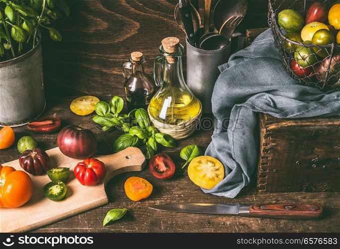 Colorful farm organic tomatoes with ingredients and cooking tools on dark rustic wooden kitchen table. Country style food background, still life, top view.
