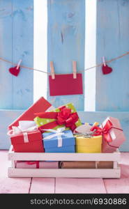 Colorful gift boxes in a white wooden crate on a pink table with a red paper note and hearts tied to a string, on a blue fence.